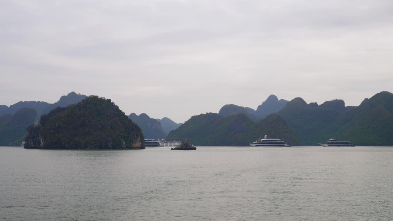 Luxury cruise ships gracefully navigate through waters of Ha Long Bay, Vietnam, surrounded by towering limestone karsts and misty mountains under an overcast sky, creating a mystical seascape