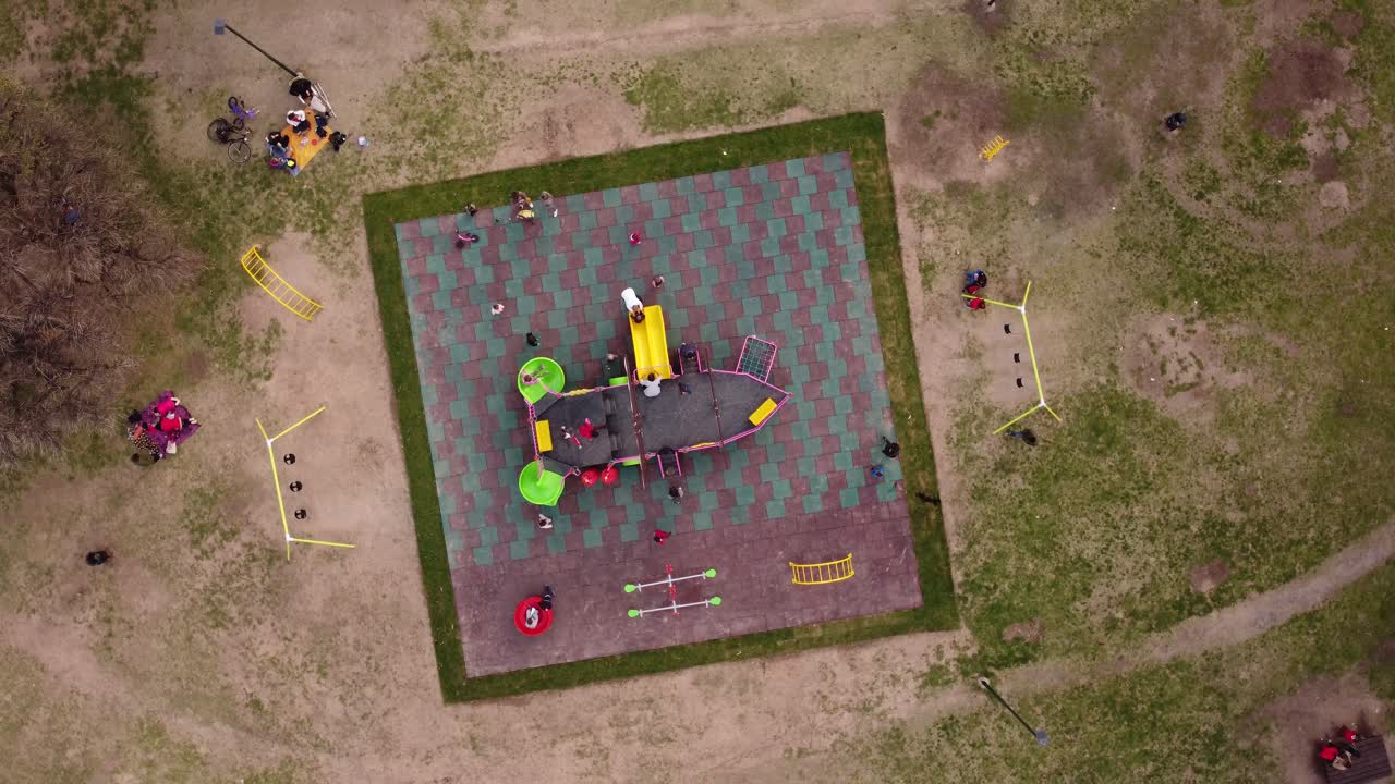 A dynamic pivoting aerial footage of kids playing around the children's playground with swings and a sailing ship-styled kiddie slide at the center