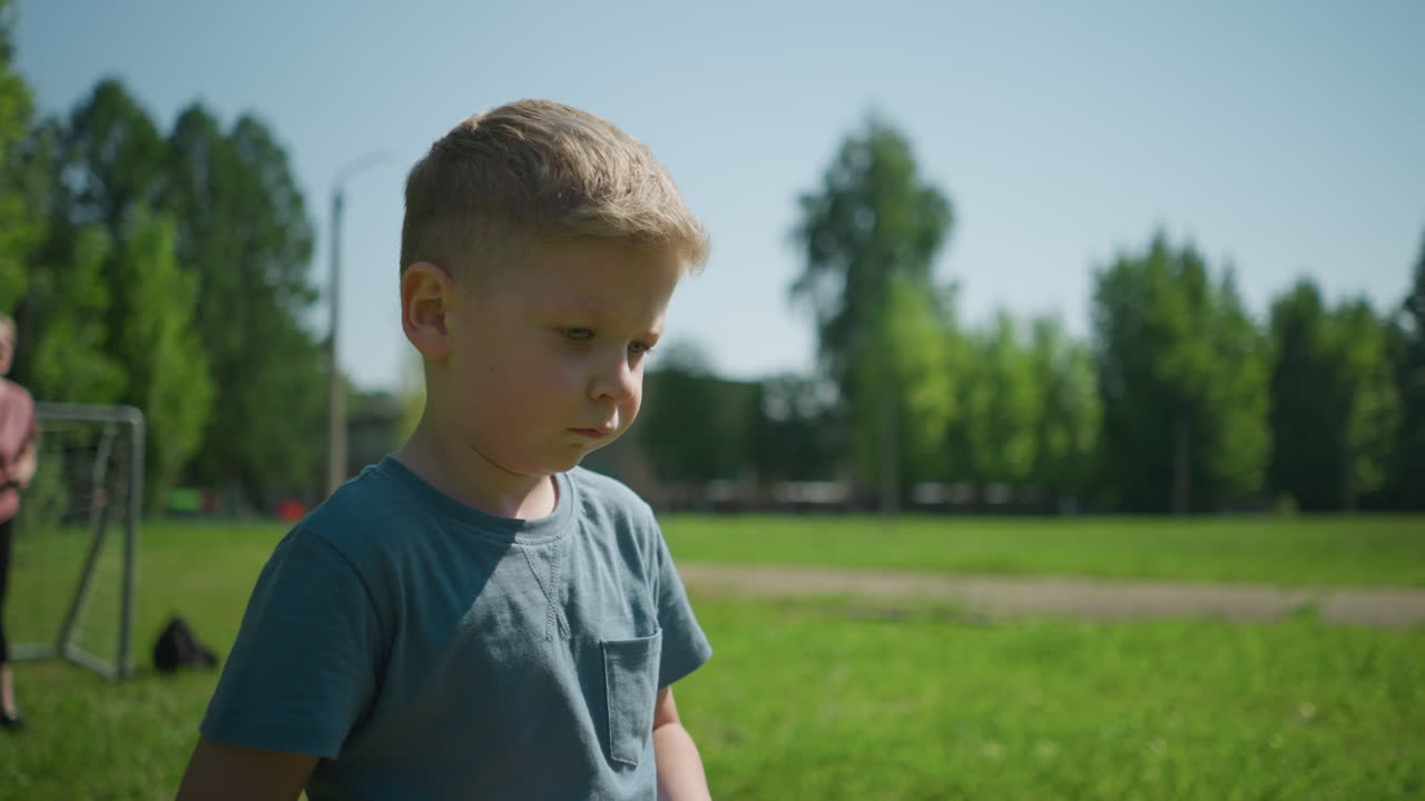 A close-up of a little child looking unhappy, with a blurred view of a woman standing in front of a goal post, set against a background of trees and a paved road in a sunny park