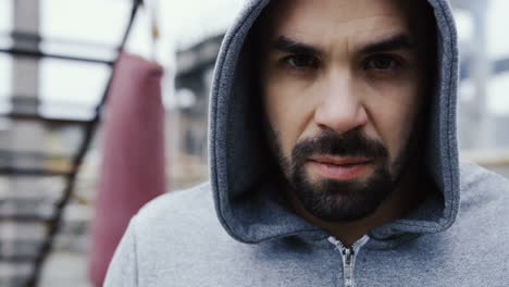 close-up view of handsome bearded man with grey hoodie and looking with serious expression at the camera outdoors an abandoned factory on a cloudy morning