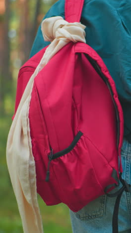 vista posterior de una mujer caminando por un bosque iluminado por el sol con una mochila roja envuelta en su hombro, ajustando la bolsa con una mano, rodeada de vegetación tranquila y luz solar filtrada