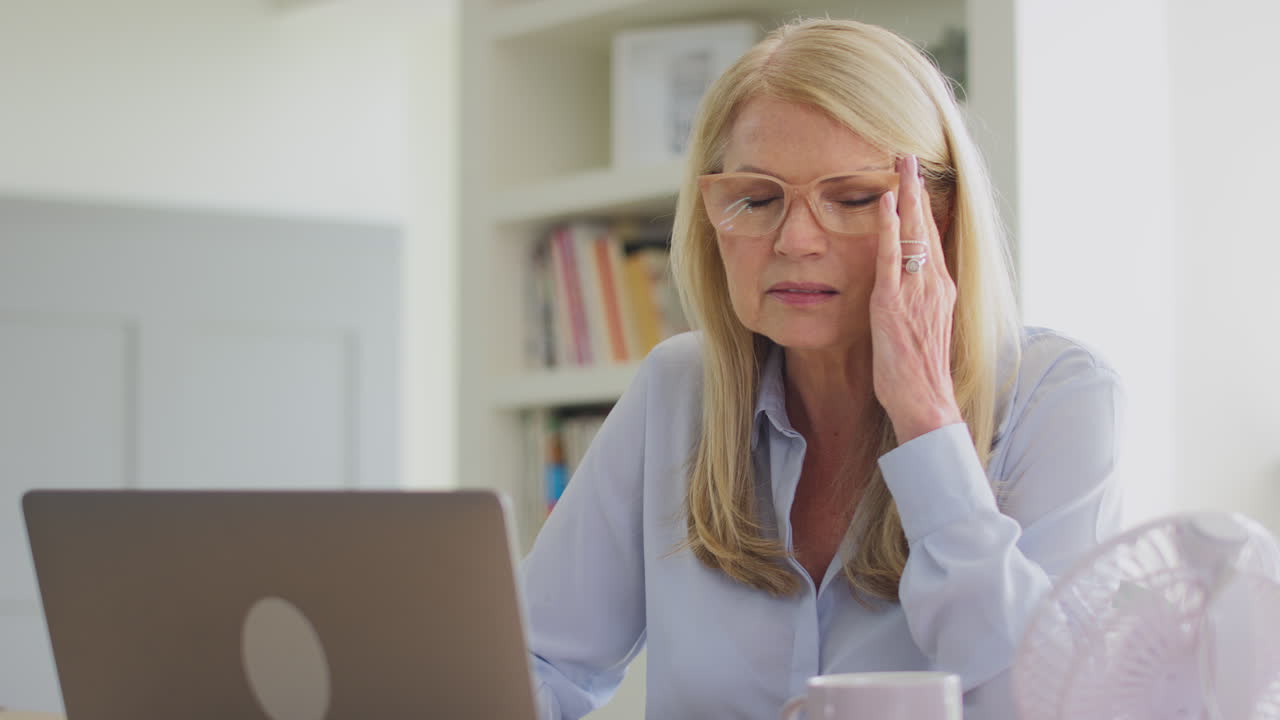 Donna matura in menopausa con gli occhiali, che lavora al laptop a casa e soffre di mal di testa