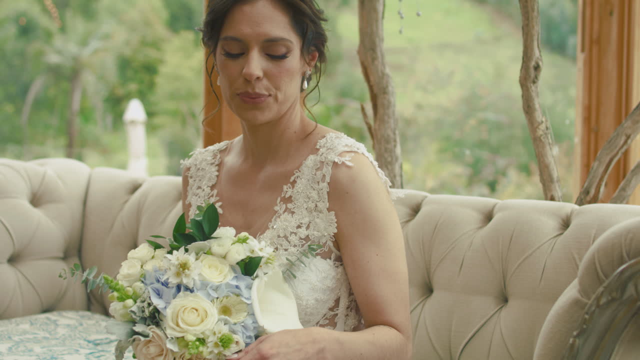 Medium shot of a bride sitting on an elegant sofa, raising her hand to look at her engagement ring. A subtle gesture full of emotion and tenderness.