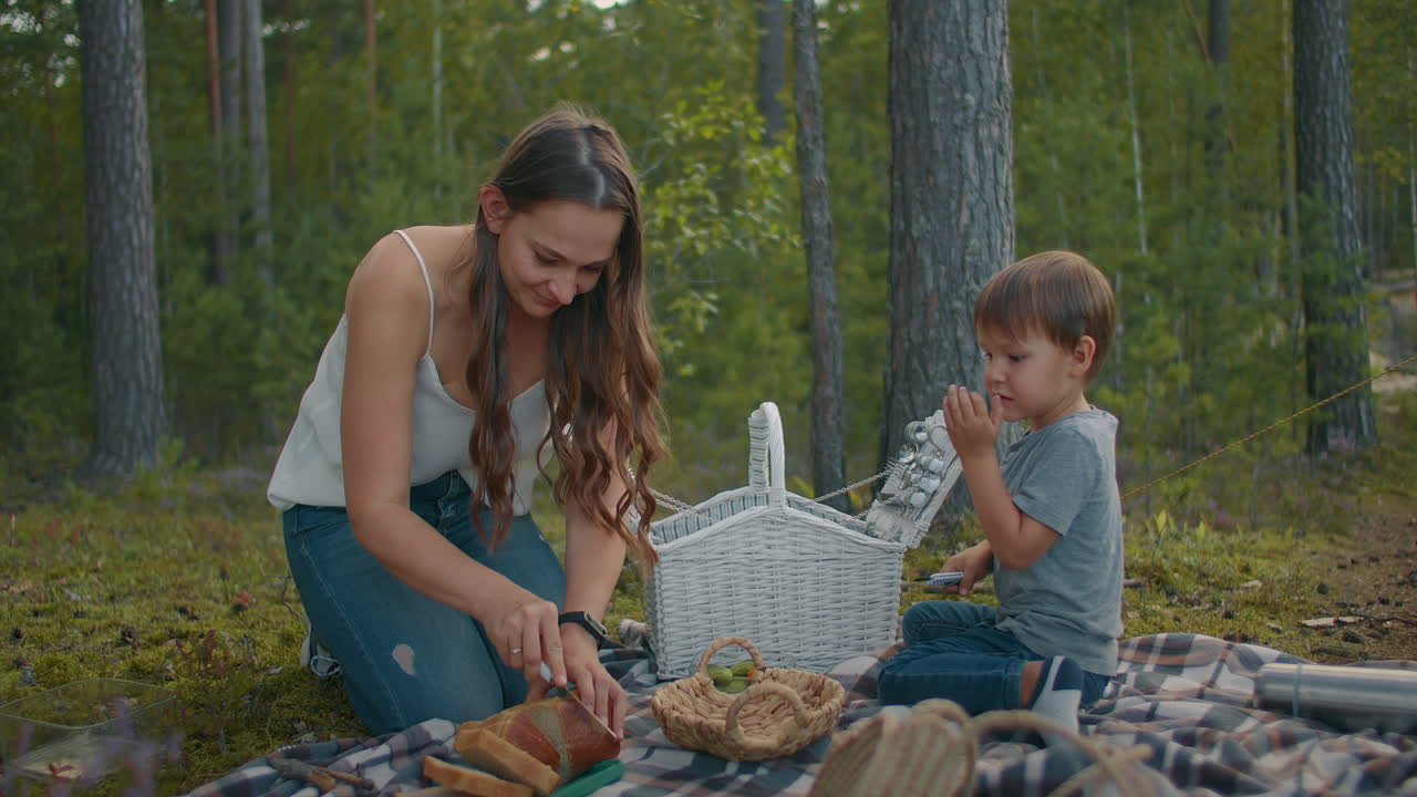 una mujer joven está cortando pan en un picnic un niño pequeño está sentado cerca de una manta descanso familiar en el bosque en verano