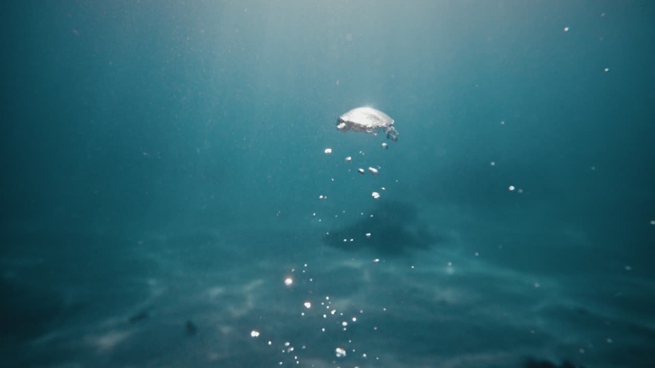 느린 움직임으로 큰 공기 거품이 깊은 푸른 푸른 물 사이로 표면으로 올라와 빛의 광선이 비추고 있습니다.