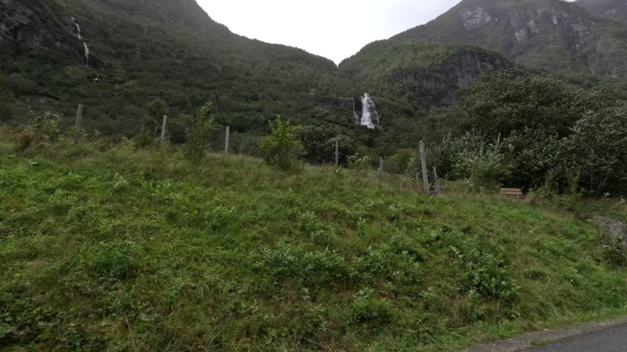 From a moving car near Flam, steep green mountains and rocky terrain frame a roadside stream and pole, capturing Norway’s dramatic fjord-valley textures and overcast atmosphere