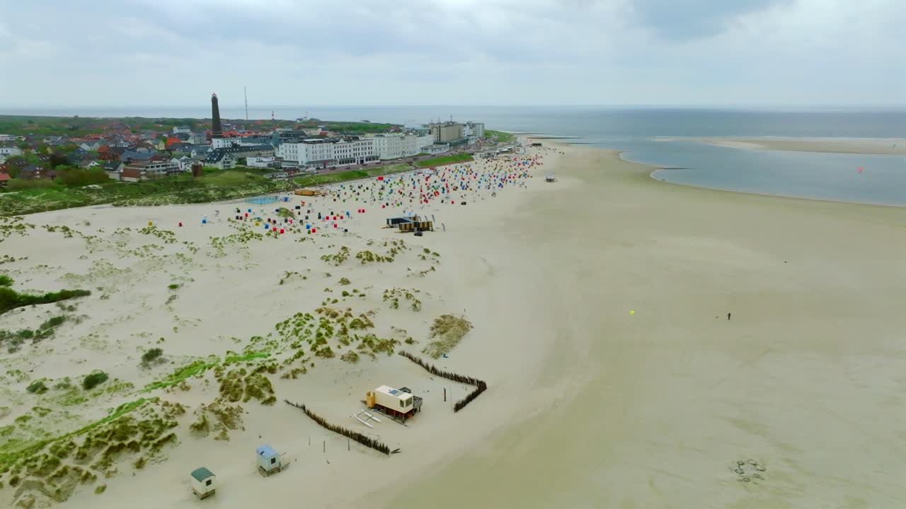 Wide sandy beach lined with colorful strandkorbe in front of Borkum town and sea. Aerial orbit view