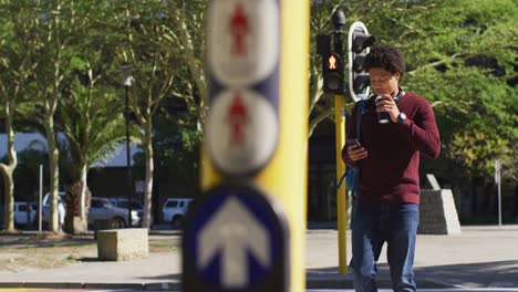 Happy-african-american-man-using-smartphone,-drinking-coffee,-crossing-street-wearing-backpack