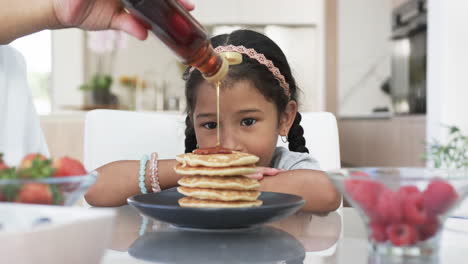 a biracial girl with dark hair smiles as syrup is poured on her pancakes