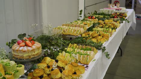 mesa llena de comida en un evento de negocios organizado dentro de almacén paredes blancas hojas de plantas colocadas en la mesa como decoración moviéndose hacia atrás revelando todos los sándwiches dulces y rollos