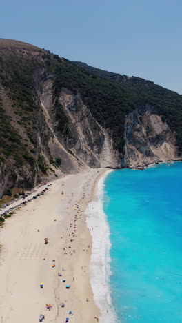 crowd of tourists at myrtos beach, cars parked at parking area, turquoise blue water with limestone mountains, kefalonia island, ionian sea, aerial, vertical shot
