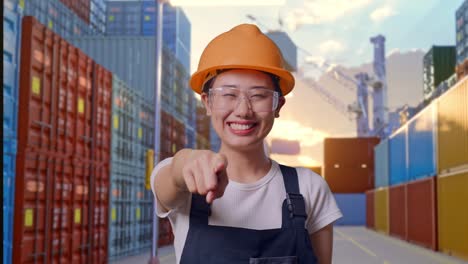 close up of asian woman worker wearing goggles and safety helmet smiling and touching her chest then pointing at you while standing at container yard warehouse
