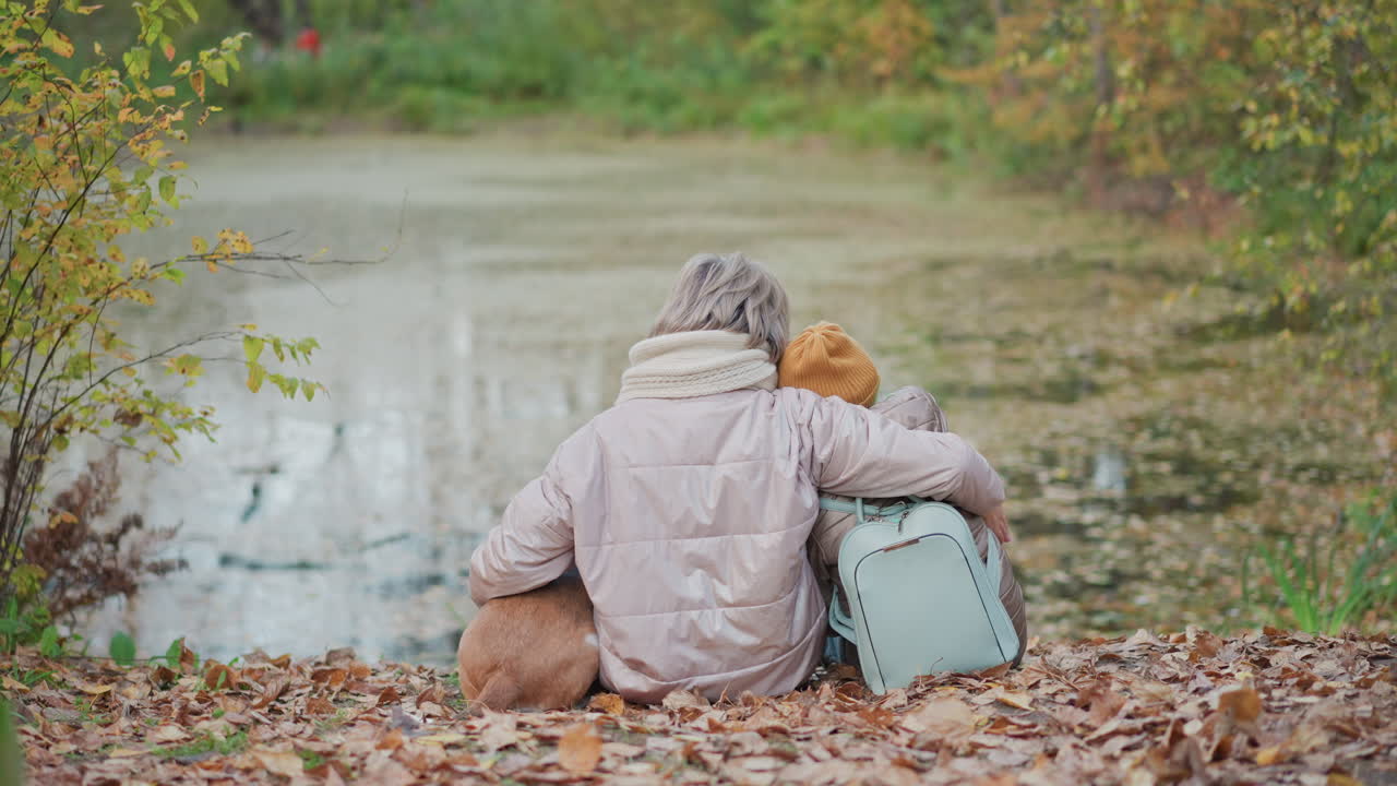 woman warmly holding dog and child close while sitting by serene leaf covered lakeside in peaceful autumn park setting, surrounded by golden foliage and quiet reflections in still water