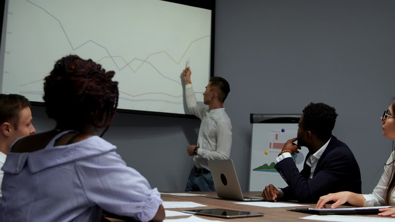 Un jeune employé de l'entreprise en chemise blanche fait un rapport sur l'avancement des travaux réalisés au cours de la dernière année et les plans de développement pour le trimestre chargé, affichés sur le diagramme du projecteur dans le bureau. Ce rapport est destiné au conseil d'administration d'une équipe multiethnique.