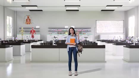 cuerpo lleno de estudiante adolescente asiática con una mochila y algunos libros sonriendo y mostrando el gesto de acuerdo mientras estaba de pie en el laboratorio de ciencias
