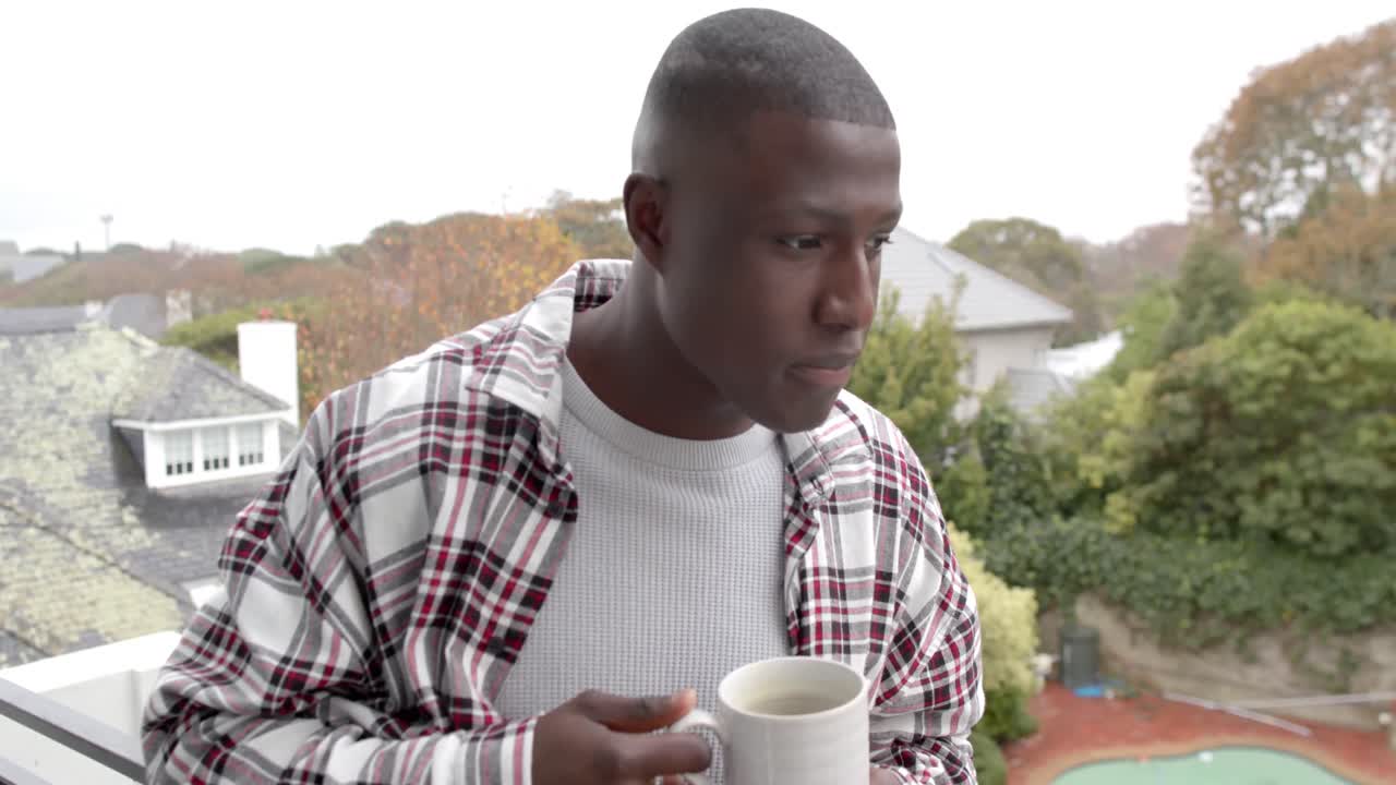 retrato de un feliz hombre afroamericano de pie en el balcón y bebiendo café en casa, cámara lenta