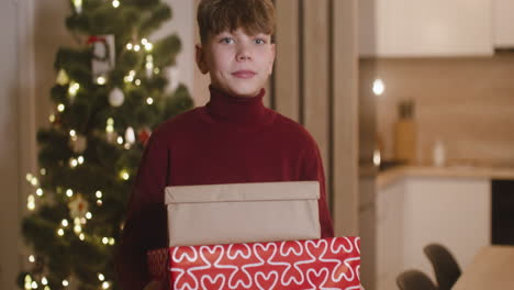 front view of a blond boy in red turtleneck sweater holding gifts in a room decorated with a christmas tree 2