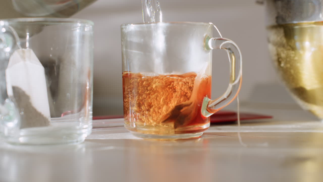 Close up of hot water being poured into glass cup with teabag inside, creating rich amber color as tea steeps beside mixing bowl in bright kitchen setting during homemade drink preparation
