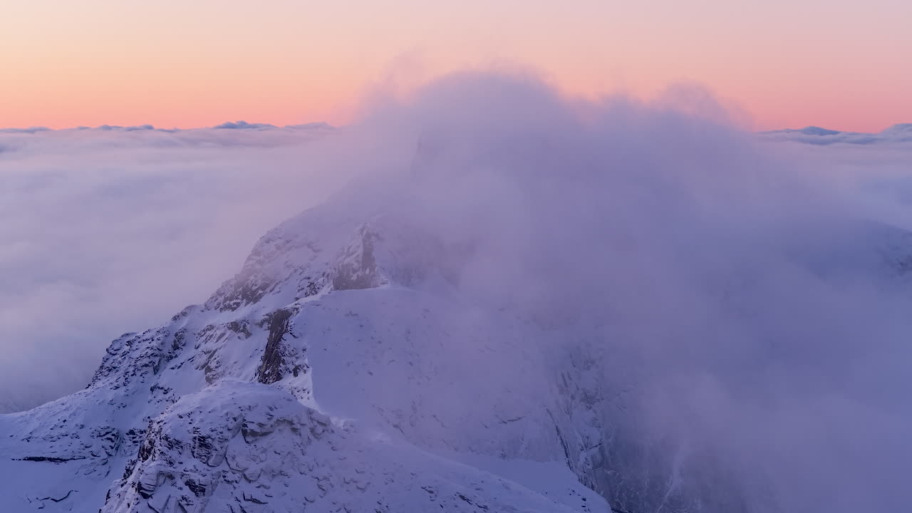 Betagende droneoptagelse af sneklædte tinder i Lofoten, Norge, der bryder frem gennem et tykt skydække under en lyserød solnedgang. Fantastisk til rejse- og naturdokumentarer.