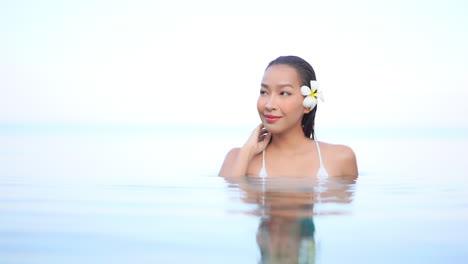 High-Key-Shot-Einer-Schönen-Jungen-Lächelnden-Asiatischen-Frau-Mit-Blume-Im-Haar,-Die-Sich-Im-überlaufpool-Entspannt