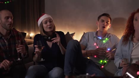 Close-up-footage-of-best-friends-celebrating,-having-time-together-at-home.-Happy-friends-sit-around-on-sofa-and-listen-to-guy-singing-and-playing-guitar.-Singing-together.-Muffled-light,-garland-lights.-Slow-motion