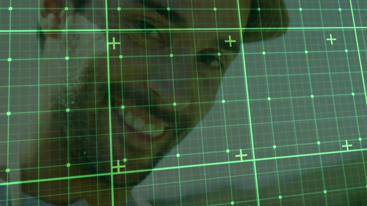 Homem inclinando a cabeça e sorrindo através de uma grade verde translúcida, reagindo a um comando na tela em um laboratório de tecnologia.