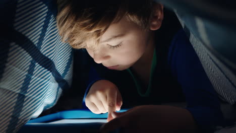 happy little boy using digital tablet computer under blanket enjoying learning on touchscreen technology playing games having fun at bedtime