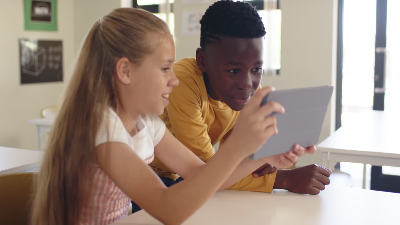 School children holding gray tablet in classroom, girl angling while boy leaning in, seeing screen