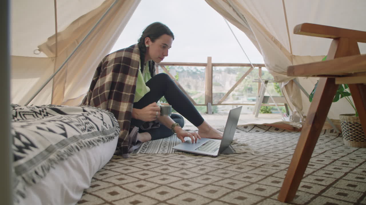 Ragazza seduta in una tenda glamping, che usa il laptop e beve tè
