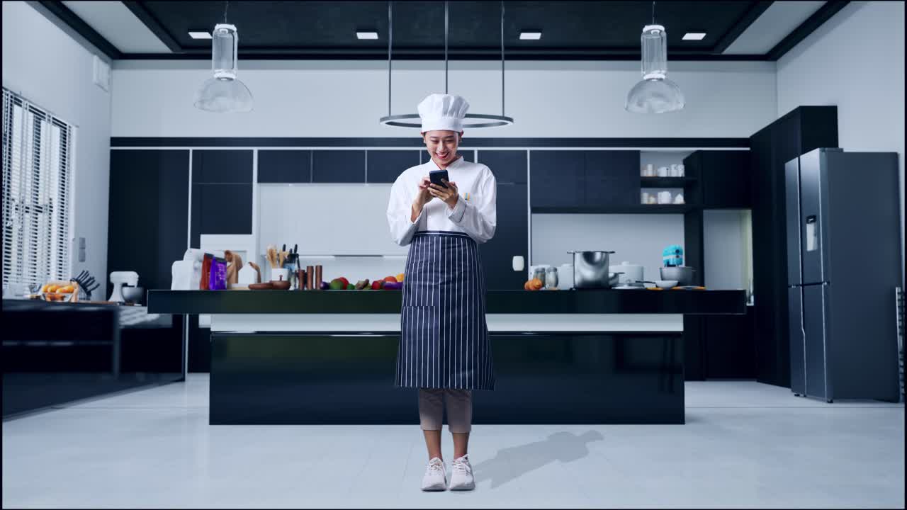 cuerpo lleno de mujer asiática chef sonriendo y señalando al teléfono inteligente en su mano en la cocina de casa