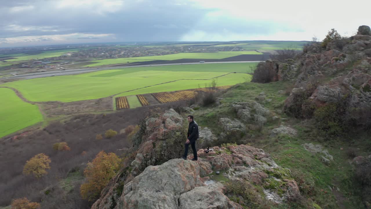 Flygfoto av en man som går till kanten av en bergsklippa och blickar ut över ett vidsträckt grönt fält, omgiven av träd i höstfärger