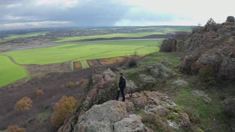 vista aérea de un hombre caminando hasta el borde de un acantilado de montaña mirando hacia un vasto campo verde, rodeado de árboles en colores de otoño