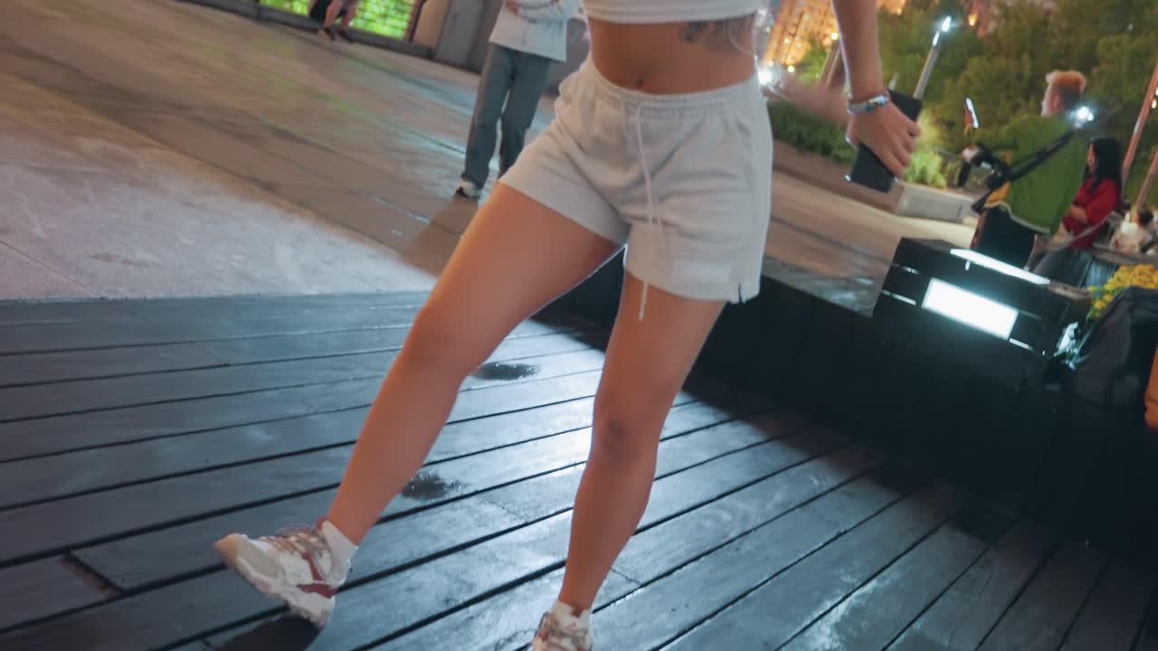 Donna asiatica ballerina in una piazza illuminata al neon, posa fiduciosa, ritratto in stile vlog e inquadrature sui passi di danza, mani dietro la testa in allungamento, pantaloncini casual e scarpe da ginnastica, auricolari, bokeh dello skyline e luci scintillanti, sera.