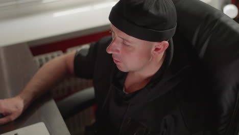 a man dressed in black is sitting at a desk in a recording studio, focused on working at a computer. the setting includes sound equipment, with another person blurred in the background