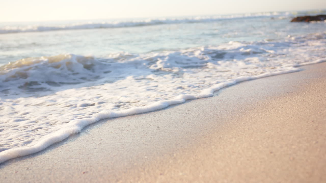 Foamy waves gently kiss the sandy beach, with sunlight reflecting off the water, with copy space