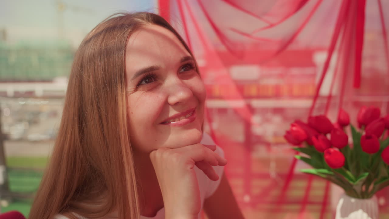 mujer caucásica en una cita romántica en una cafetería rodeada de una cortina roja y un ramo de tulipanes, sonrisa alegre y mirada espontánea hacia la ventana, luz suave y horizonte de la ciudad visible, ambiente íntimo y cálido