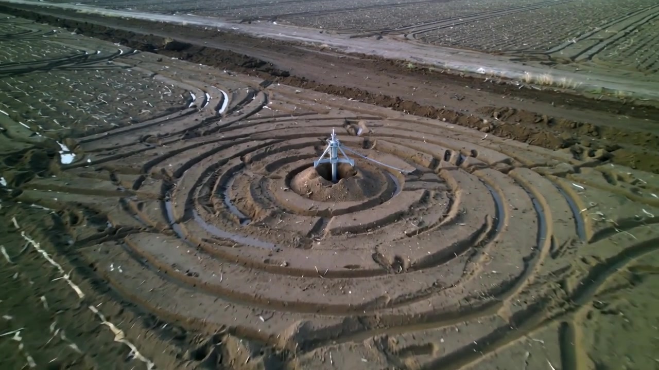 Approaching drone inspecting metal irrigation head on low mound in tilled field, circular ruts