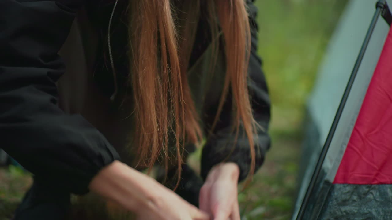 close up of lady hand gripping thick stick to pull metal tent peg from ground near forest tent as pine needles scatter on soil and red tent corner remains anchored beside camping gear