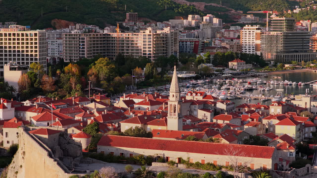 Aerial drone view of Budva Old Town, showing its medieval stone buildings, red rooftops, fortress walls, and church tower along the coastline, with modern city buildings and mountains in the background. Montenegro