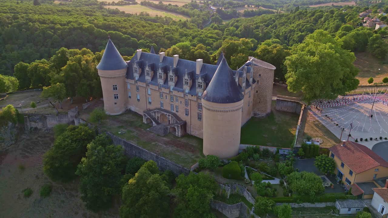 Drone pullback shot at sunset showing the iconic Rochechouart Castle, home to the Contemporary Art Museum of Haute-Vienne, with surrounding village, forest, and fields - France