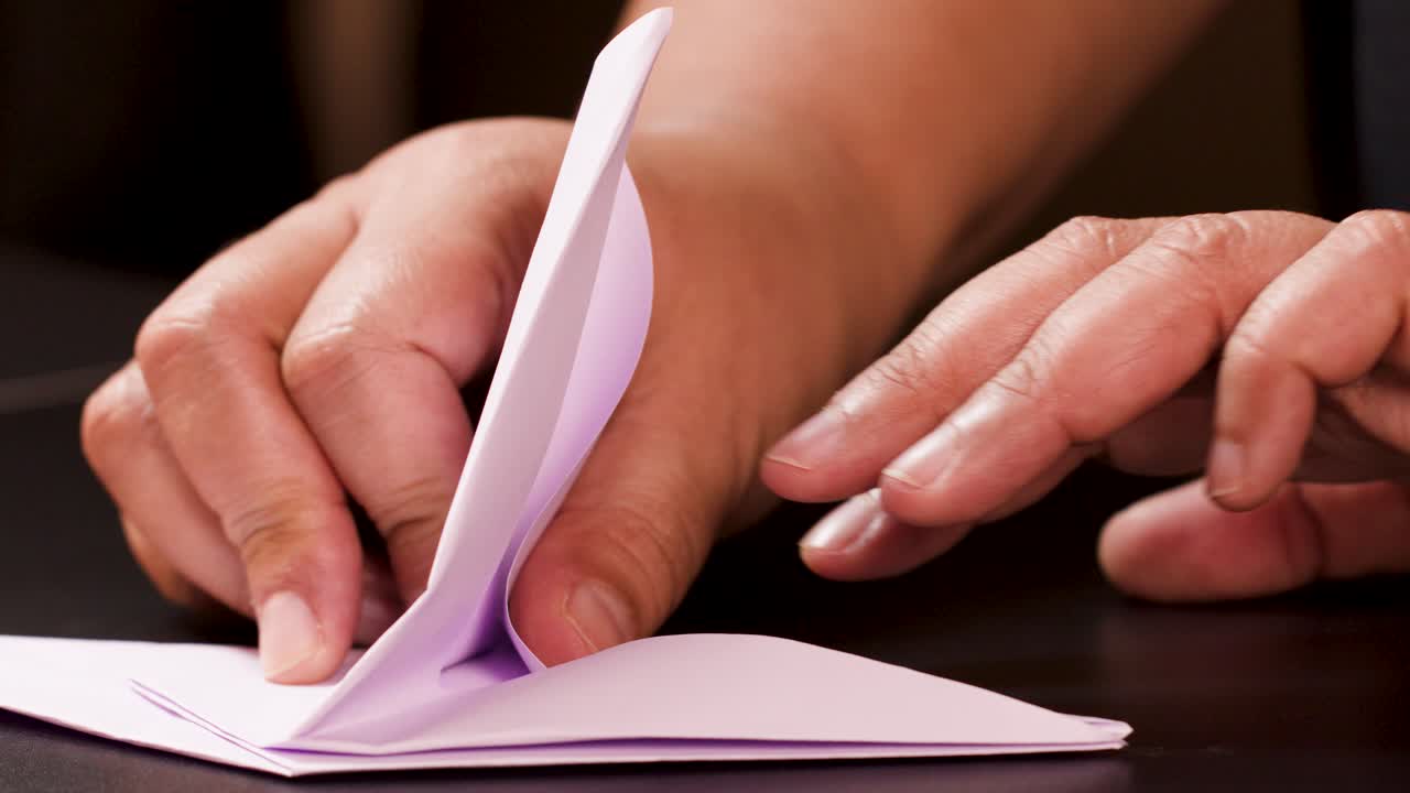 Adult hands carefully fold white paper into an origami boat, close-up, warm lighting, steady camera