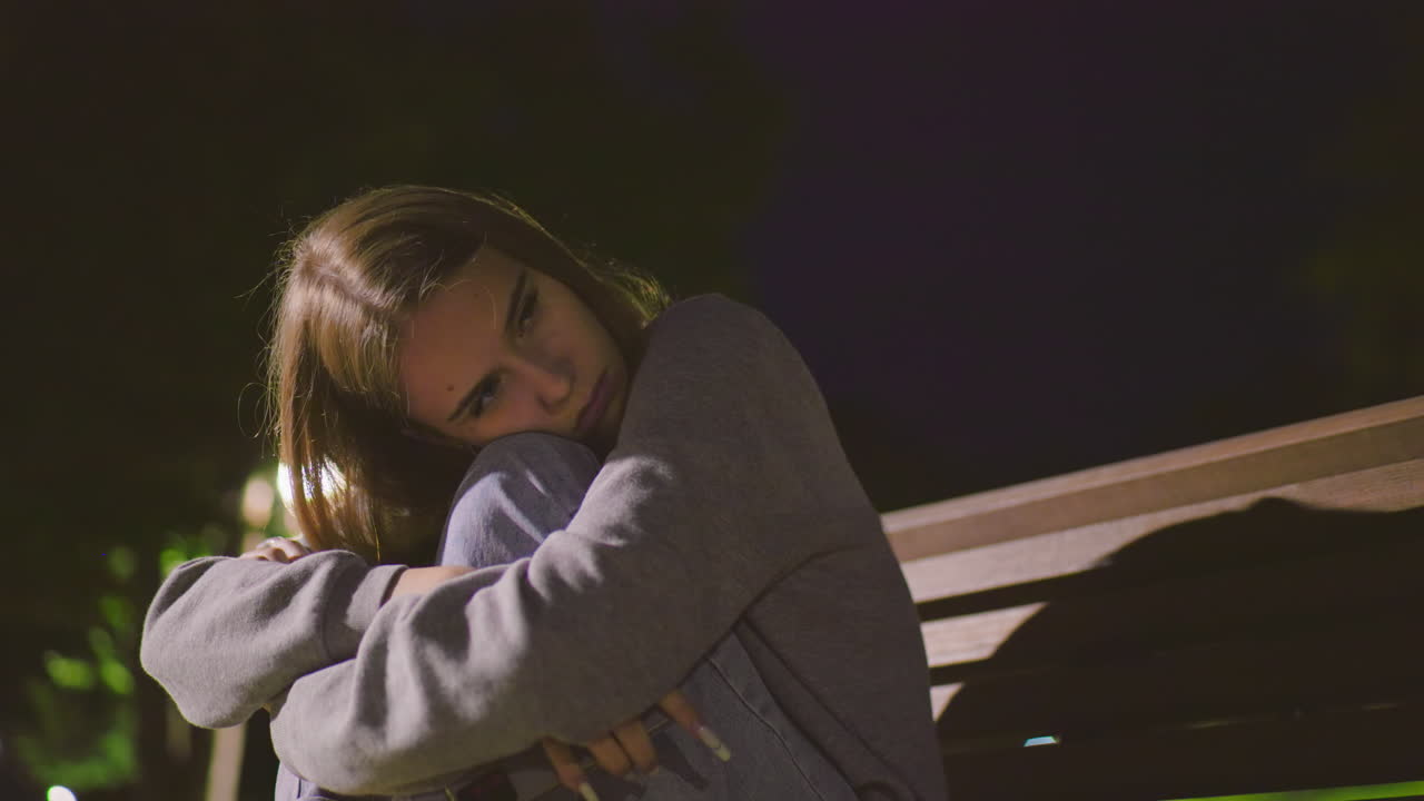 dama sentada en un banco del parque por la noche con los brazos cruzados sobre las rodillas, descansando la cabeza pensativamente, mirando a la distancia bajo la suave iluminación del parque, expresando calma contemplación y estado de ánimo reflexivo