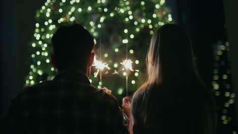 A-young-couple-holds-burning-sparklers-in-their-hands,-sits-facing-a-large-Christmas-tree-at-home