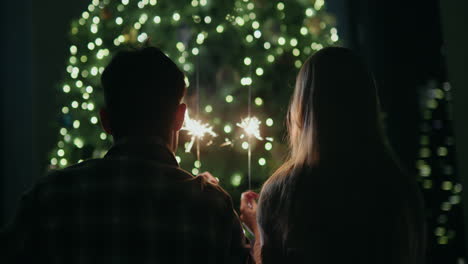 Una-Pareja-Joven-Sostiene-Bengalas-Encendidas-En-Sus-Manos,-Se-Sienta-Frente-A-Un-Gran-árbol-De-Navidad-En-Casa