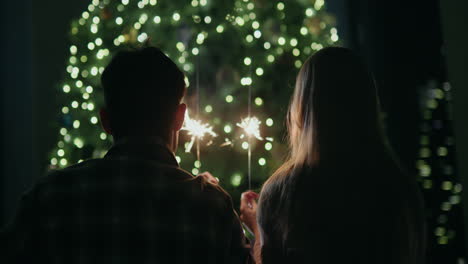 una pareja joven sostiene chispas ardientes en sus manos, se sienta frente a un gran árbol de navidad en casa