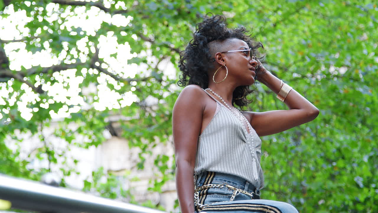 Trendy young black woman wearing camisole, side stripe jeans and sunglasses, sitting on handrail playing with her hair, low angle