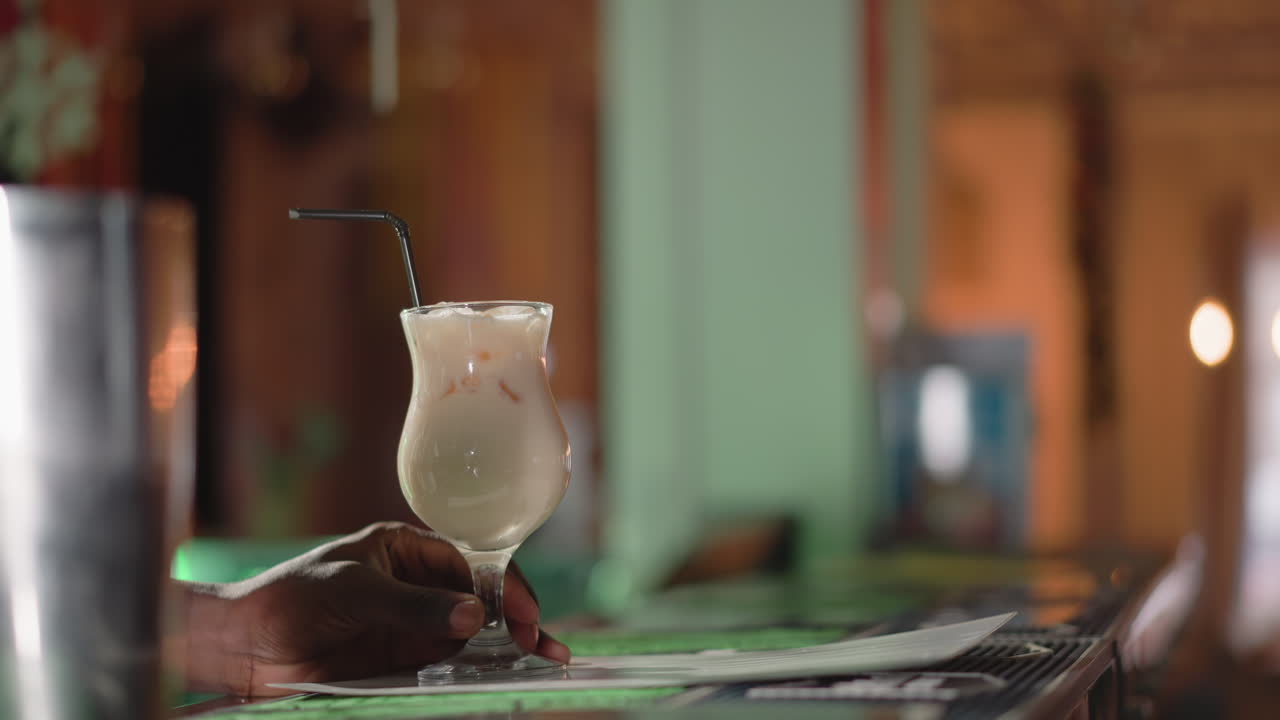 Bartender stirring creamy drink with straw in glass on bar counter, focused on hand, drink, and smooth blending of ingredients in a relaxed bar setting