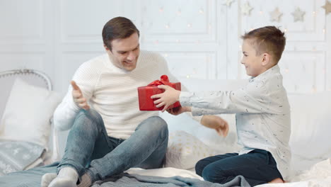 Surprised-father-getting-new-year-present-in-bedroom.-Happy-son-presenting-gift