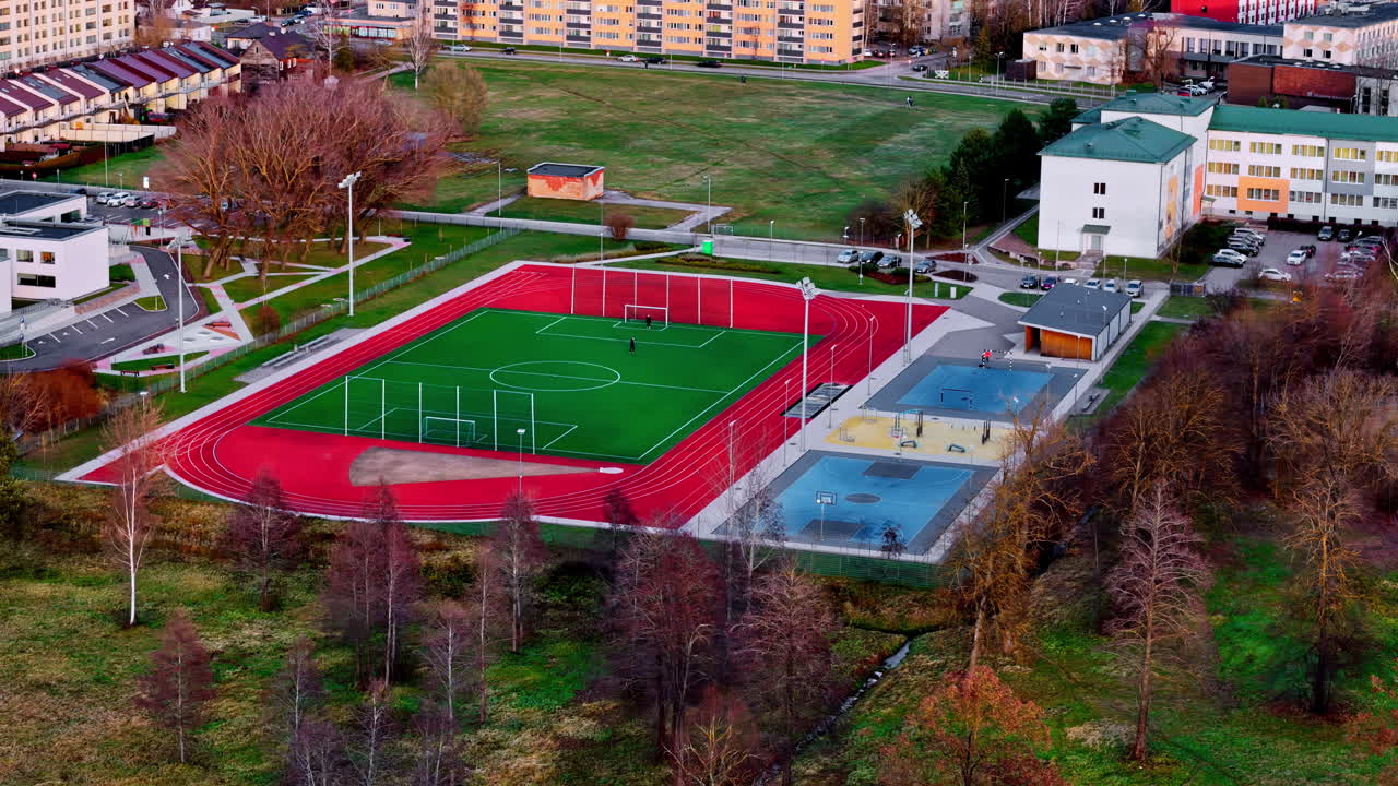 Two people playing football on a vibrant red sports field surrounded by trees and buildings in autumn