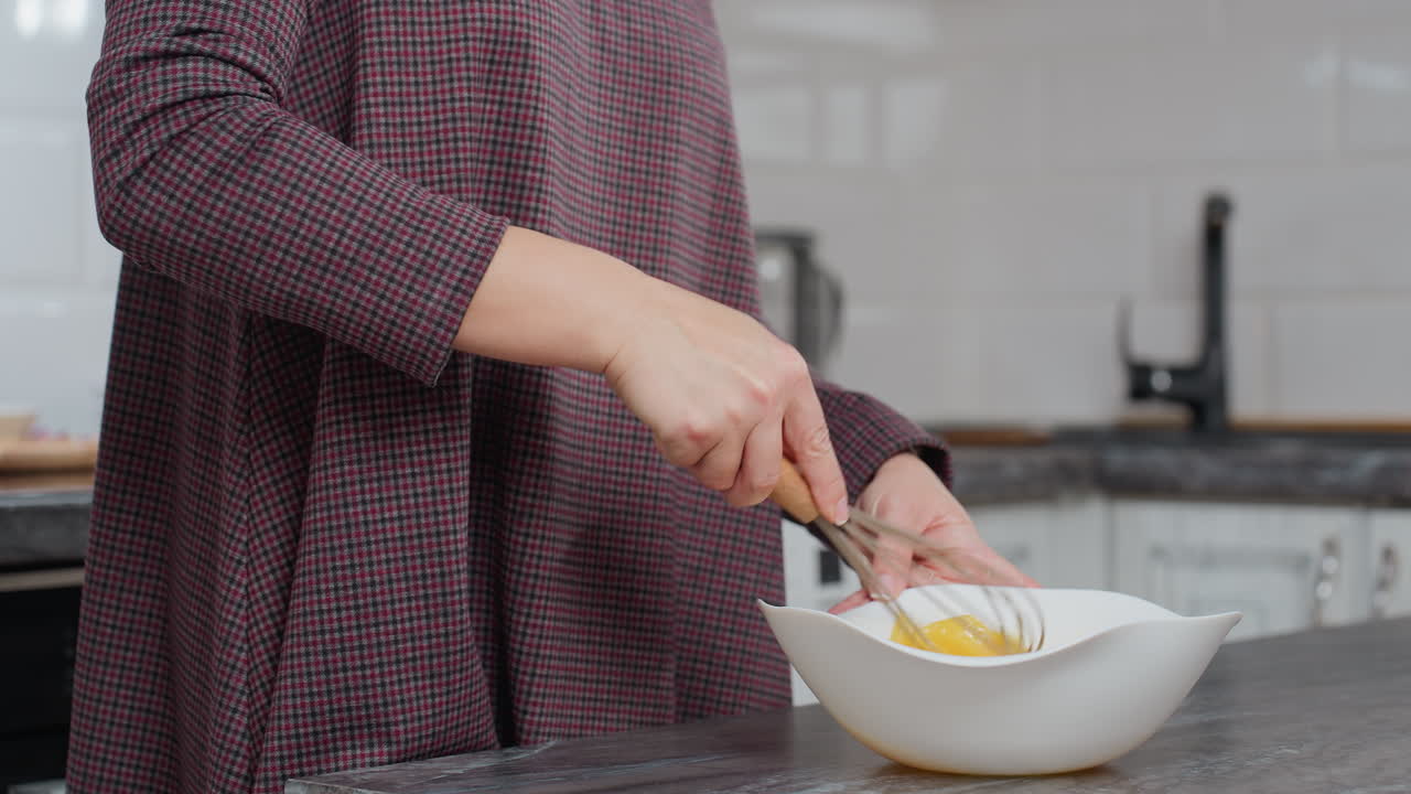 vista parcial de una mujer con túnica de damas agitando huevo en un cuenco blanco mientras se prepara para hornear, el fondo borroso muestra equipos de cocina, utensilios y interiores modernos